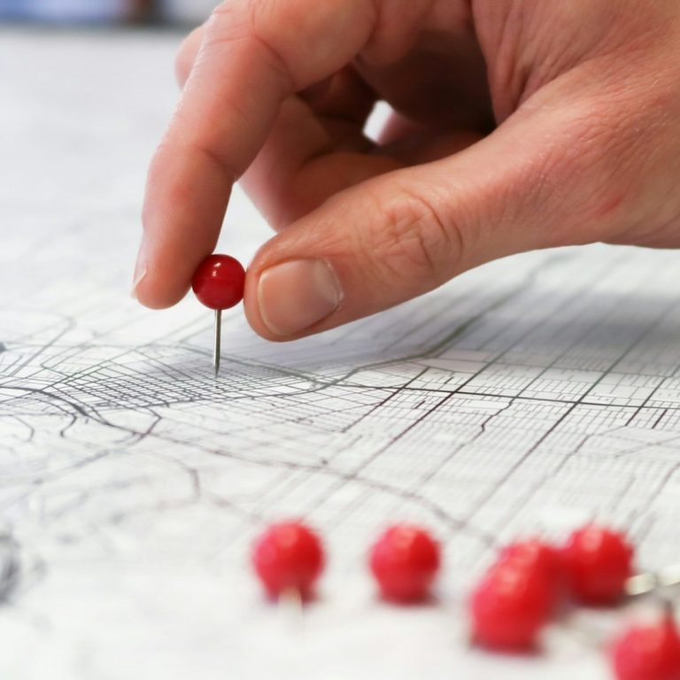 immer auf den Punkt genaue Planung Eine Hand platziert eine rote Stecknadel auf einer Landkarte. Weitere Stecknadeln liegen daneben.